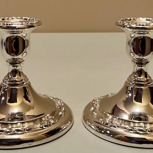 May include: A pair of polished silver-colored metal candle holders featuring a wide, circular base with a decorative beaded edge. Each holder has a smooth, tapered stem and a flared top rim designed to securely hold a standard taper candle.