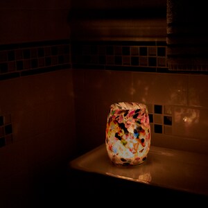 May include: A colorful glass candle holder with a white background. The candle holder is lit and casting a warm glow.