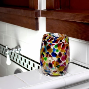 May include: A colorful glass candle holder with a speckled design in shades of red, orange, yellow, green, blue, and purple. The holder is sitting on a white tiled surface.