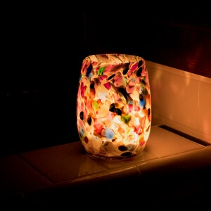 May include: A colorful glass candle holder with a white background and multi-colored spots. The candle holder is lit and glowing.