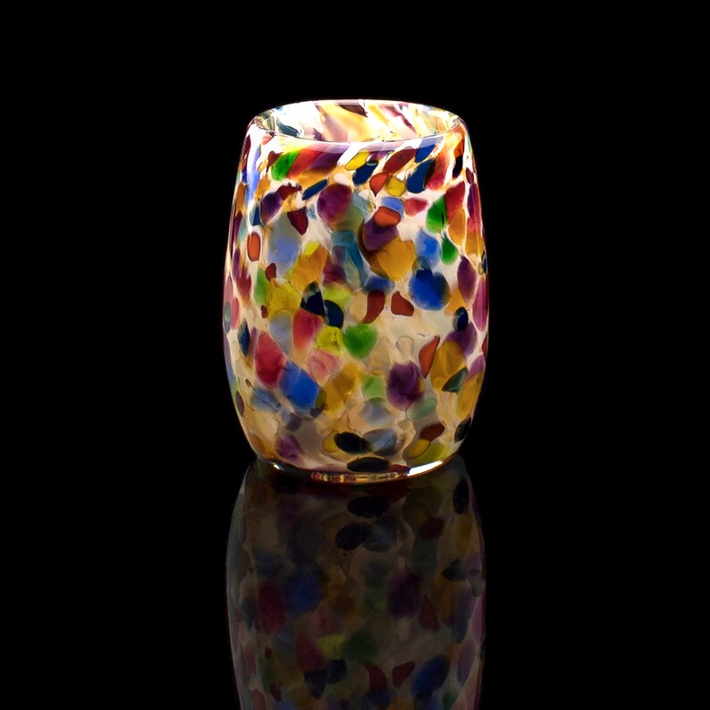 May include: A colorful glass tumbler with a speckled pattern in shades of red, blue, green, yellow, and purple. The tumbler is made of clear glass with a slightly opaque finish.