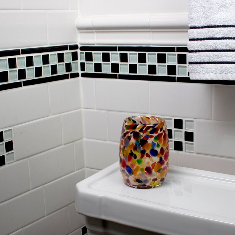 May include: A colorful glass candle holder with a speckled design sits on a white bathroom counter. The counter is tiled with white subway tiles and a black and white checkerboard border.