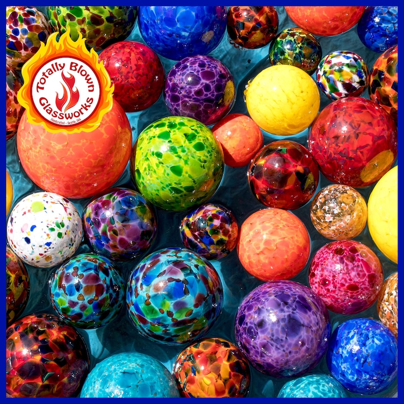 May include: A collection of colorful glass orbs with speckled patterns, floating in a blue pool of water. The orbs are various sizes and shapes, with colors including red, orange, yellow, green, blue, purple, and white. The image includes the logo "Totally Blown Glassworks".