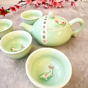 Puede incluir: Un juego de té de cerámica verde celadón con una tetera y varias tazas pequeñas. Cada taza tiene una figura de pez koi en su interior. La tetera tiene una tapa con un pequeño pez koi y un asa. El juego se exhibe con flores rosas.