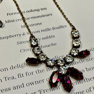 May include: A vintage necklace with a fine chain and a pendant adorned with clear and deep red gemstones. The pendant has a floral design. The necklace is set against a backdrop of printed text.