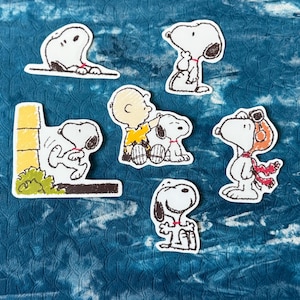 Peut inclure: Une collection de six autocollants de personnages Snoopy dans diverses poses. Les autocollants présentent Snoopy dans différentes tenues et activités, avec un fond blanc et des contours noirs. Les autocollants sont sur un fond bleu.