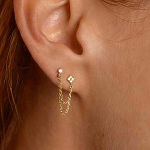 May include: A pair of gold earrings with a chain connecting two studs. One stud is a small round diamond, and the other is a four-petal flower design with a diamond center. The earrings are worn on a person's ear.