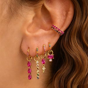 Puede incluir: Pendientes y puño de oreja dorados con piedras preciosas de colores. Los pendientes incluyen una variedad de diseños, como gotas, círculos y formas rectangulares, todos adornados con piedras rosas, verdes, azules y amarillas. El puño de oreja es una doble banda de gemas rosas y moradas.