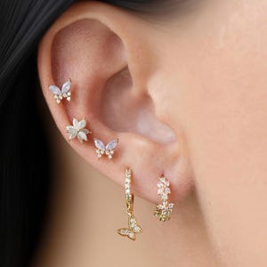 May include: A collection of gold-toned earrings, including butterfly and floral designs, adorn an ear. The earrings feature small, sparkling gemstones. The butterfly earrings dangle from the ear, adding a touch of elegance.