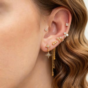 May include: A collection of gold-toned earrings adorns an ear. The earrings include small studs, starburst designs, and dangling chain earrings. Some earrings are embellished with small, clear gemstones. The ear is framed by wavy, light brown hair.