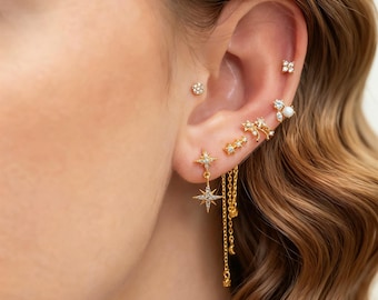 Könnte beinhalten: Ein Ohr ist mit einer Auswahl an goldfarbenen Ohrringen geschmückt. Die Ohrringe umfassen kleine Ohrstecker, sternförmige Designs und hängende Kettenohrringe. Einige Ohrringe sind mit kleinen, klaren Edelsteinen verziert. Das Ohr wird von welligem, hellbraunem Haar umrahmt.