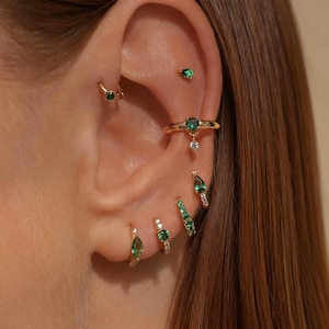 May include: An assortment of gold earrings and ear cuffs, all featuring emerald green gemstones. The earrings include studs, hoops, and a dangling ear cuff, presenting a coordinated jewellery set for multiple ear piercings.