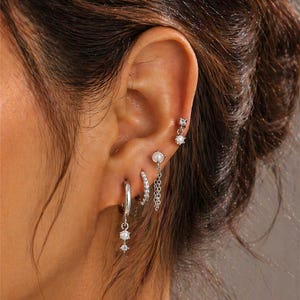 Può includere: Una collezione di orecchini d'argento adorna un orecchio. Gli orecchini includono piccoli borchie, cerchi con ciondoli pendenti e un orecchino a catena. Gli orecchini presentano pietre preziose trasparenti e perle, creando un aspetto delicato ed elegante.
