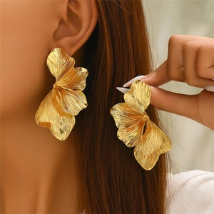 Puede incluir: Pendientes dorados con un diseño texturizado en forma de pétalo. Los pendientes tienen una forma en capas, similar a un abanico, y se muestran en primer plano. Una mano sostiene los pendientes.