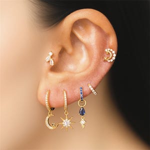 May include: A close-up of an ear adorned with multiple gold earrings. The earrings feature celestial designs like moons and stars, and some are embellished with clear and blue gemstones. The jewelry is set against the backdrop of the ear and skin.