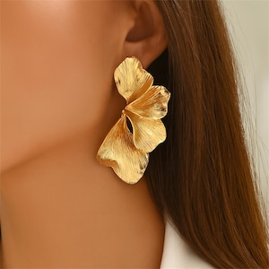 May include: Gold-colored statement earrings. Each earring features a layered, petal-like design with textured detailing. The earrings are worn on a person's ear, showcasing their size and shape. The earrings have a shiny, metallic finish.