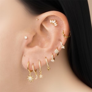 May include: A collection of gold earrings and ear piercings. The earrings include hoops, studs, and dangling designs with stars, moons, and geometric shapes. The jewelry is adorned with small, sparkling gemstones.