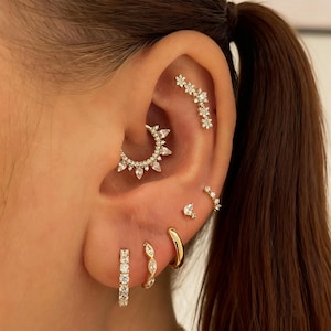 May include: An ear adorned with multiple pieces of gold-toned jewelry. The pieces include a hoop earring, a small stud, a curved bar with small gems, a floral-shaped piece, and a larger hoop with pointed gems.