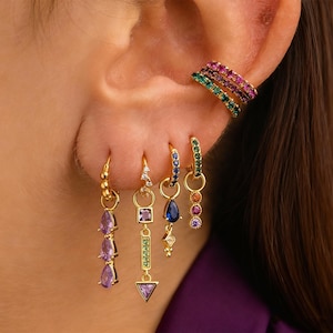 May include: Gold-toned earrings with colorful gemstones. The earrings include hoop styles and dangling designs with purple, blue, green, and pink stones. A matching ear cuff with pink and green stones is also visible.