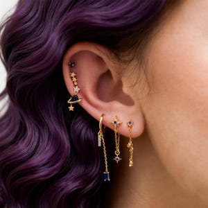 May include: Gold earrings with a celestial theme. The earrings feature star, planet, and chain designs with blue gemstone accents. The ear piercings are displayed against a backdrop of purple hair.