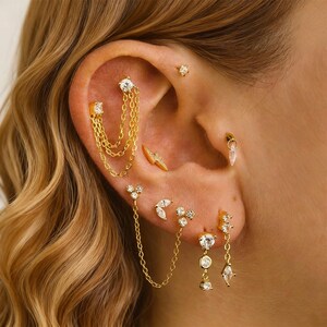 Op de afbeelding: Een close-up van een oor versierd met meerdere gouden oorbellen. De oorbellen hebben verschillende ontwerpen, waaronder studs, kettinghangers en stukken met heldere edelstenen.