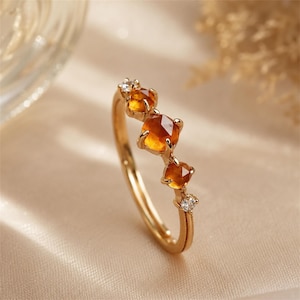 Puede incluir: Un anillo de oro con tres piedras preciosas de color naranja-amarillo y dos pequeñas piedras blancas. El anillo tiene una banda delicada. Las piedras son cuadradas y en forma de corazón. Fondo neutro.