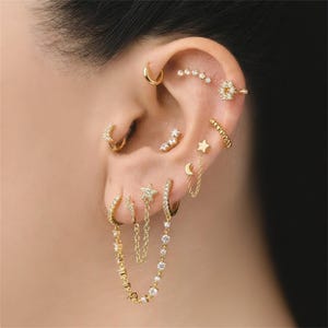 May include: An ear is adorned with multiple gold-coloured earrings. The collection includes hoops, studs, and chain designs. Some earrings are set with small, sparkling stones, while others feature star and crescent moon shapes.