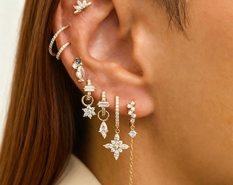 Könnte beinhalten: Ein Ohr mit mehreren goldenen Ohrringen. Die Ohrringe weisen verschiedene Designs auf, darunter kleine Stecker, Creolen und hängende Stücke mit klaren Edelsteinen. Oben am Ohr befindet sich ein kleiner goldener Stecker mit floralem Design.