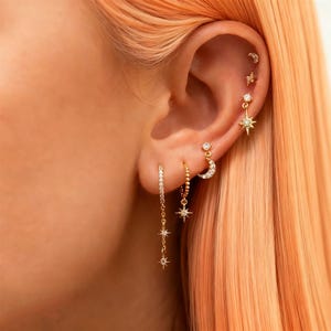 May include: A close-up of an ear displaying several gold earrings. The earrings include a dangling chain with three stars, a hoop with a star pendant, a crescent moon with a star, and several small studs. The earrings are decorated with small, sparkling stones.