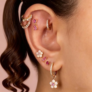 Puede incluir: Una colección de pendientes dorados con piedras preciosas rosas y blancas. Los pendientes incluyen aros, tachuelas y un diseño floral colgante. La oreja se muestra sobre un fondo rosa suave, mostrando los detalles de la joyería.