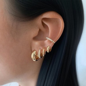 Peut inclure: Ensemble de boucles d'oreilles dorées et un ear cuff assorti. Les boucles d'oreilles comprennent trois petits anneaux et deux anneaux plus grands, dont un avec de petites pierres claires. L'ear cuff est orné de petites pierres claires.