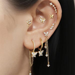 May include: A collection of gold-colored earrings and ear piercings, featuring butterfly and leaf designs. The earrings include hoops and dangling chains with sparkling accents. The piercings are set with clear gemstones.