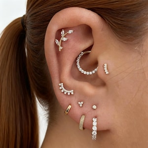 Può includere: Primo piano di un orecchio adornato con più gioielli. L'orecchio presenta una varietà di orecchini d'oro, tra cui un orecchino a forma di foglia, un cerchio con pietre trasparenti e diversi piccoli borchie e cerchi. I gioielli sono appoggiati sulla pelle.