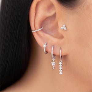 May include: A close-up of an ear adorned with multiple pieces of jewelry. A silver ear cuff, a small cluster stud, and three different styles of earrings are visible. The earrings include small hoops and dangling designs, all featuring clear, sparkling stones.