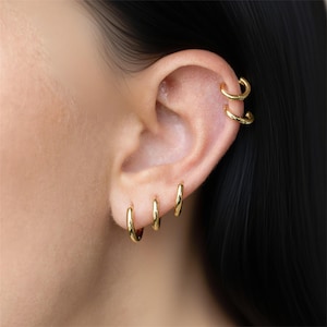 May include: Close-up of an ear with multiple gold hoop earrings. Three small hoops are in the earlobe, and two larger hoops are in the upper ear cartilage. The earrings have a polished, shiny finish.