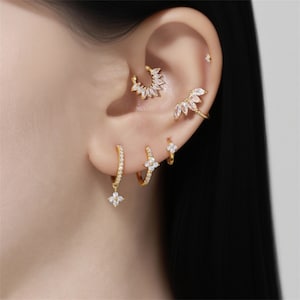 Può includere: Una collezione di orecchini e polsini dorati, adornati con pietre preziose chiare e scintillanti. I gioielli includono orecchini a cerchio con ciondoli floreali, piccoli cerchietti e un polsino decorativo. I pezzi sono disposti su un orecchio.