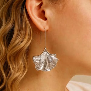 Può includere: Orecchini pendenti color argento con design a foglia. Gli orecchini hanno la forma di una foglia di ginkgo con venature dettagliate. Sono appesi a un sottile gancio di filo. Lo sfondo mostra un orecchio e una spalla.