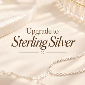 Puede incluir: Un collar de cadena de plata sobre una superficie clara con el texto marrón oscuro Upgrade to Sterling Silver centrado sobre un pequeño icono de corazón. Los eslabones metálicos de la cadena son visibles en primer plano con luz suave.