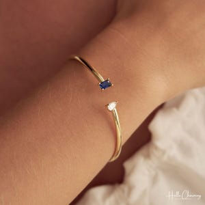 Puede incluir: Una pulsera abierta de oro con una piedra preciosa de zafiro azul rectangular en un extremo y una piedra blanca transparente en forma de pera en el otro. La banda de metal pulido es delgada y circular, diseñada para la muñeca.