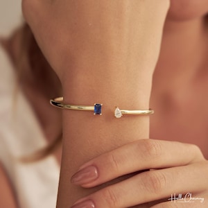 May include: A gold-toned open cuff bracelet featuring a rectangular blue gemstone on one end and a pear-shaped clear gemstone on the other. The jewellery piece has a smooth, polished finish and is positioned on a wrist. The text Hello Charming is visible.
