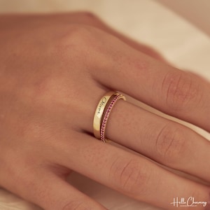 Peut inclure: Une bague double en ton doré composée d'un anneau lisse gravé du prénom Oliver et d'un second anneau plus fin serti d'une rangée de petites pierres roses éclatantes. Les deux anneaux sont reliés pour former un seul bijou.