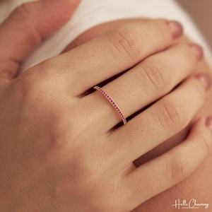 May include: A thin gold band ring featuring a continuous row of small, round, vibrant pink gemstones set along the outer surface. The ring has a polished metallic finish and a delicate, slender profile that encircles the finger with uniform stones.