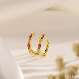 May include: A pair of gold hoop earrings featuring alternating marquise-cut red stones and round light pink stones. The jewelry displays the text Hello Charming in the bottom right corner. The hoops have a polished finish and a hinged closure.