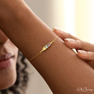 May include: A gold chain bracelet featuring a pea pod-shaped centerpiece containing three small, round, multi-colored gemstones in yellow, blue, and purple. The delicate metal chain connects to the central pod design, creating a refined jewelry piece.