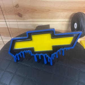 May include: A yellow, black, and blue Chevrolet logo with a dripping paint effect. The logo is a stylized bow tie shape with a blue outline and blue drips. The background is a black surface.