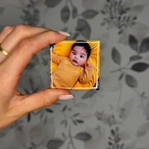 K&ouml;nnte beinhalten: Ein kleiner quadratischer Magnet mit einem Foto eines Babys mit dunklen Haaren, das ein senfgelbes Langarmshirt tr&auml;gt. Das Baby liegt auf einem leuchtend gelben Stoff. Das Foto ist auf der Magnetoberfl&auml;che mit einem d&uuml;nnen wei&szlig;en Rand versehen.