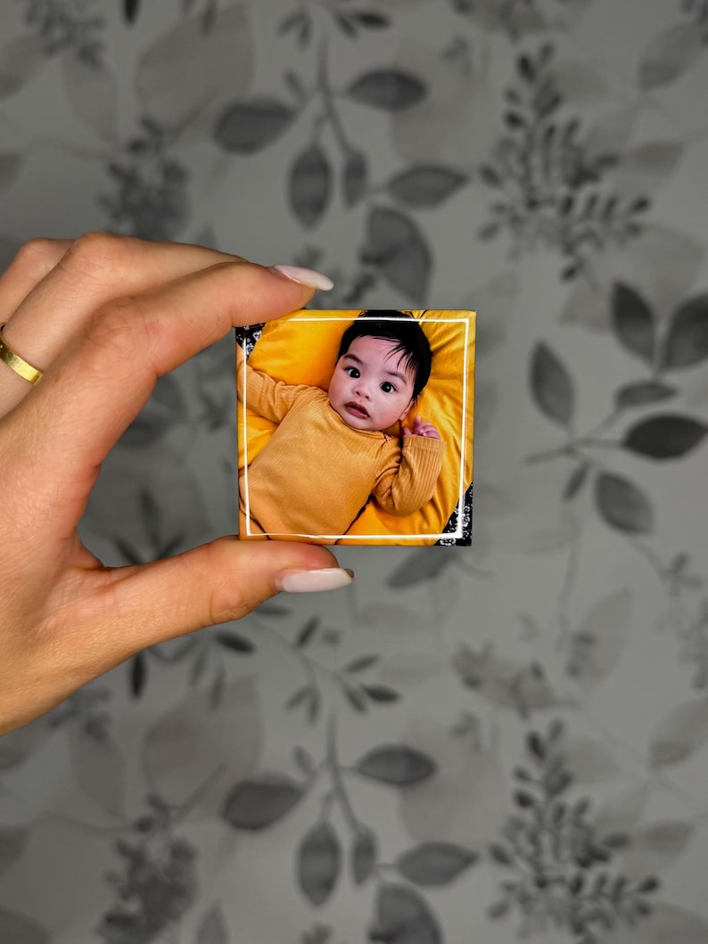 K&ouml;nnte beinhalten: Ein kleiner quadratischer Magnet mit einem Foto eines Babys mit dunklen Haaren, das ein senfgelbes Langarmshirt tr&auml;gt. Das Baby liegt auf einem leuchtend gelben Stoff. Das Foto ist auf der Magnetoberfl&auml;che mit einem d&uuml;nnen wei&szlig;en Rand versehen.