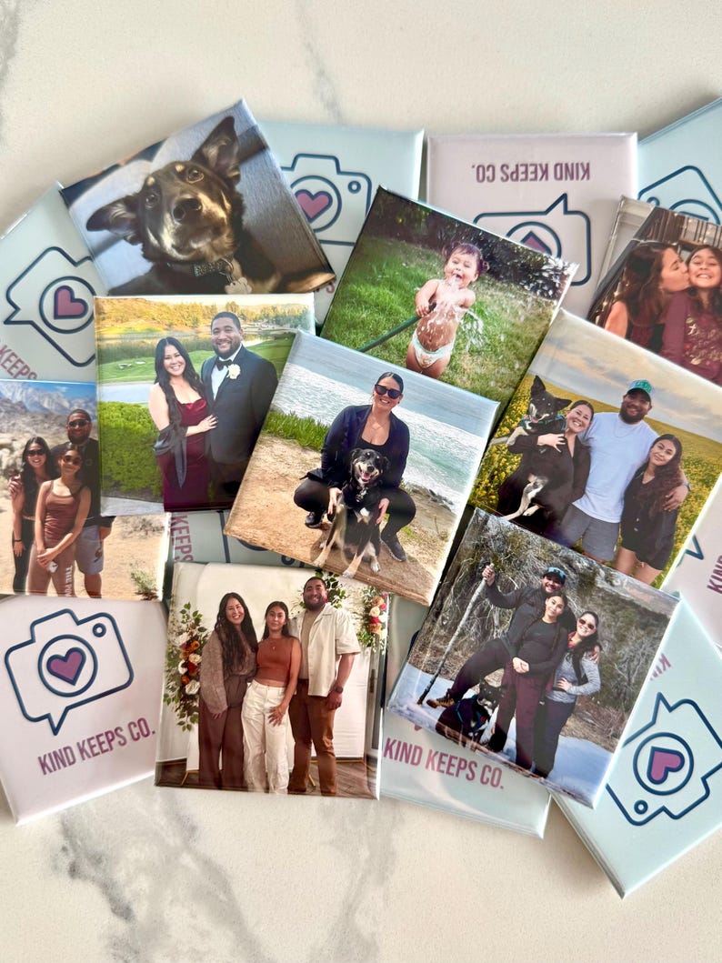 K&ouml;nnte beinhalten: Eine Sammlung quadratischer Magnete mit verschiedenen gedruckten Fotos von Menschen, Hunden und Kindern sowie hellblauen Magneten, die ein Kamerasymbol mit einem Herz und den Text Kind Keeps Co. auf einer wei&szlig;en Oberfl&auml;che zeigen.