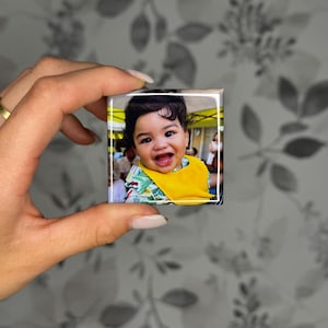 K&ouml;nnte beinhalten: Ein quadratischer Fotomagnet mit einem l&auml;chelnden Baby mit dunklem Haar, das ein leuchtend gelbes L&auml;tzchen und ein Hemd mit gr&uuml;nem Blattmuster tr&auml;gt. Das Bild ist auf eine gl&auml;nzende Oberfl&auml;che mit wei&szlig;em Rand gedruckt und zeigt eine Szene im Freien.