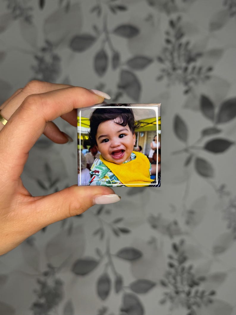 K&ouml;nnte beinhalten: Ein quadratischer Fotomagnet mit einem l&auml;chelnden Baby mit dunklem Haar, das ein leuchtend gelbes L&auml;tzchen und ein Hemd mit gr&uuml;nem Blattmuster tr&auml;gt. Das Bild ist auf eine gl&auml;nzende Oberfl&auml;che mit wei&szlig;em Rand gedruckt und zeigt eine Szene im Freien.